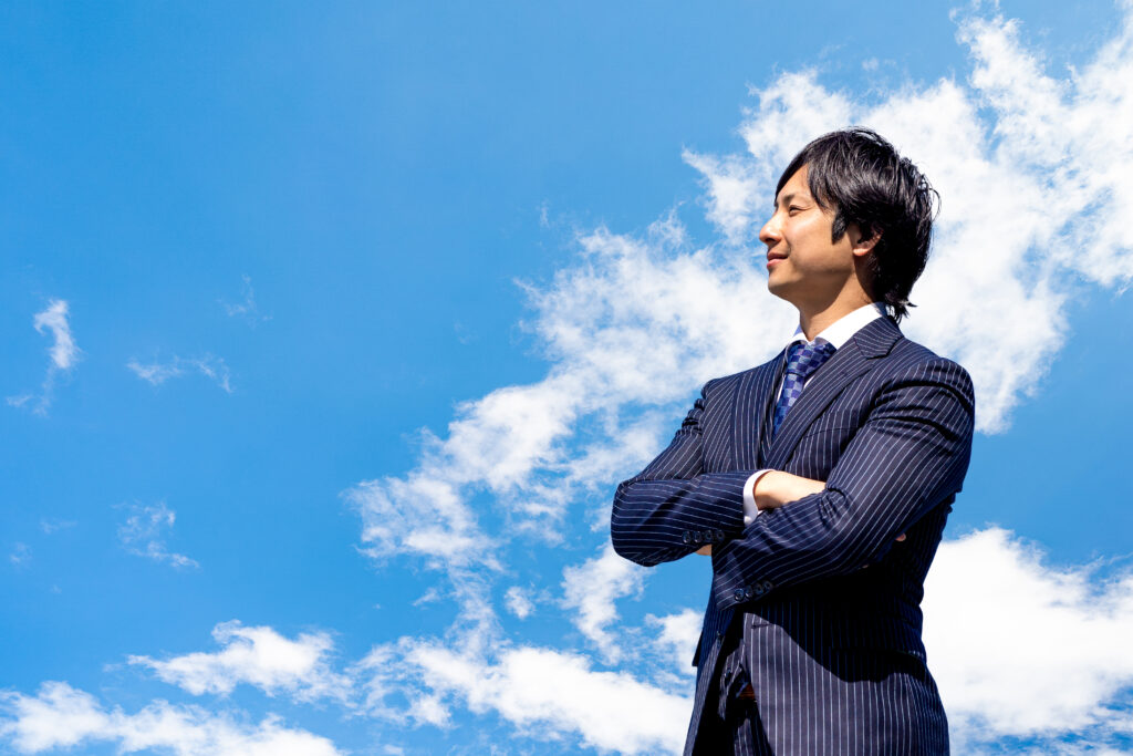 青空をバックに腕組みをするスーツの男性